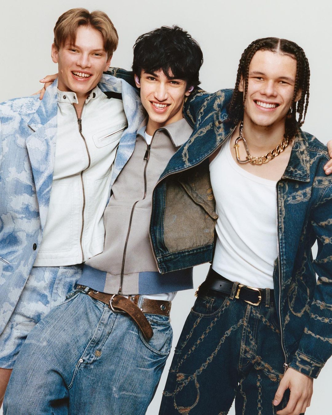Three young men pose in an embrace, showcasing modern fashion with denim jackets and jeans in a bright, clean indoor setting.
