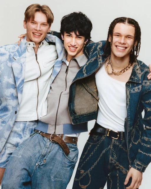 Three young men pose in an embrace, showcasing modern fashion with denim jackets and jeans in a bright, clean indoor setting.