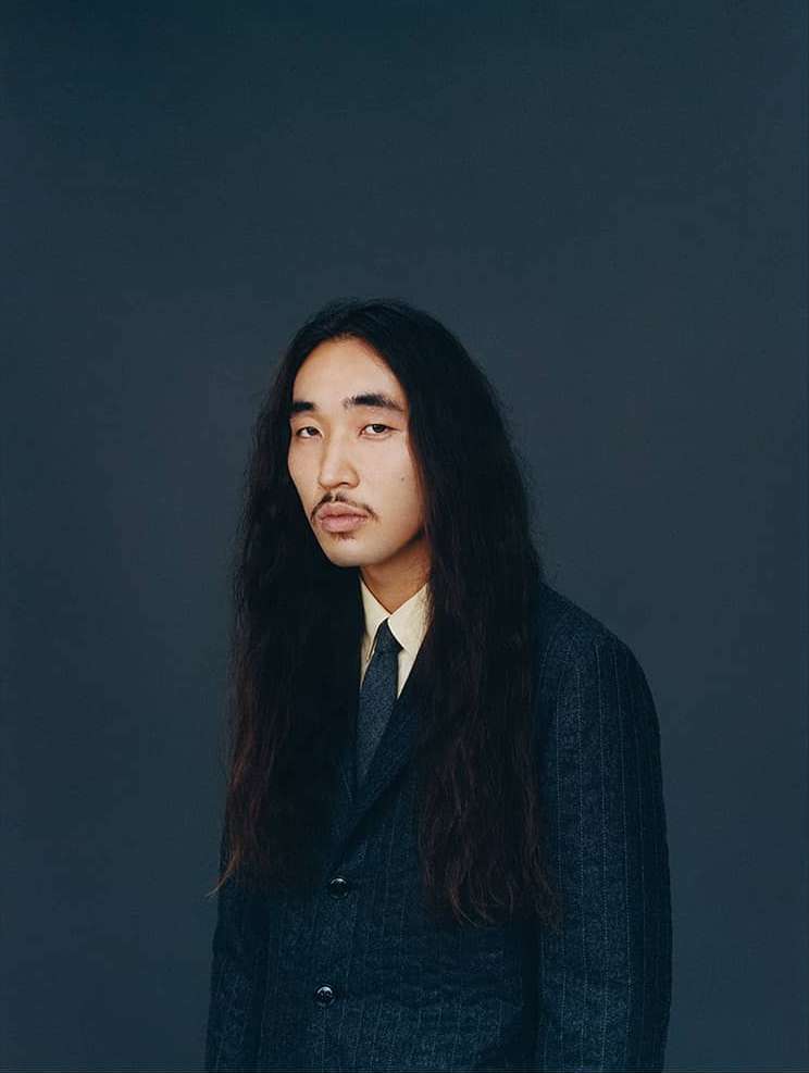 Portrait of an adult man with long dark hair, wearing a suit with a light-yellow dress shirt and tie, against a solid grey background.