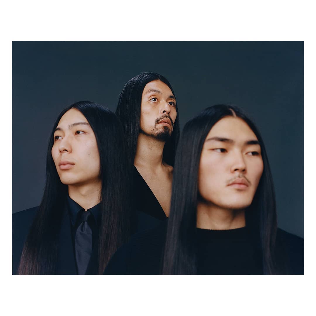 Three Asian men with long black hair pose indoors against a gray backdrop, showcasing a blend of fashion and cultural identity.