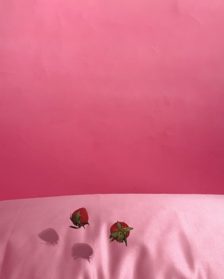 Two red strawberries with green stems are on a pink satin surface against a pink backdrop in a colorful, food-focused still life.