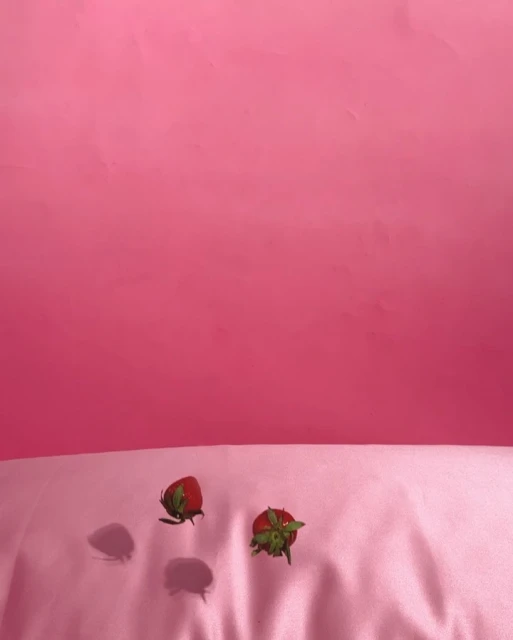 Two red strawberries with green stems are on a pink satin surface against a pink backdrop in a colorful, food-focused still life.