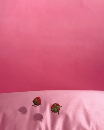 Two red strawberries with green stems are on a pink satin surface against a pink backdrop in a colorful, food-focused still life.