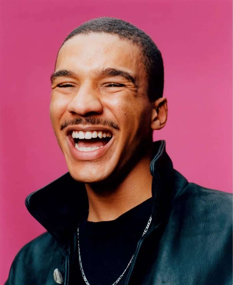 Close-up portrait of a smiling young adult male with a mustache wearing a black leather jacket against a pink background.