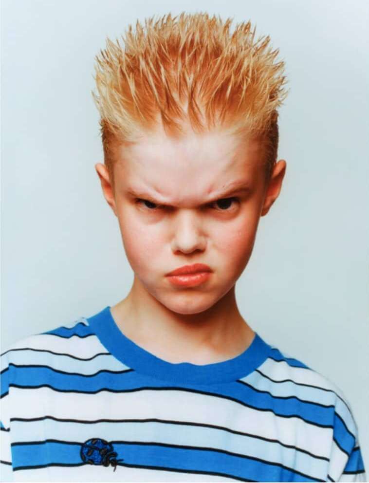 Portrait of a blonde, young boy with spiky hair and a striped t-shirt, looking at the camera with an angry facial expression.