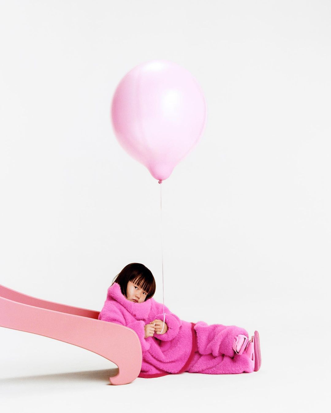 Child in a pink outfit sits on a pink slide holding a pink balloon against a white backdrop in a studio. Playful and minimalist.