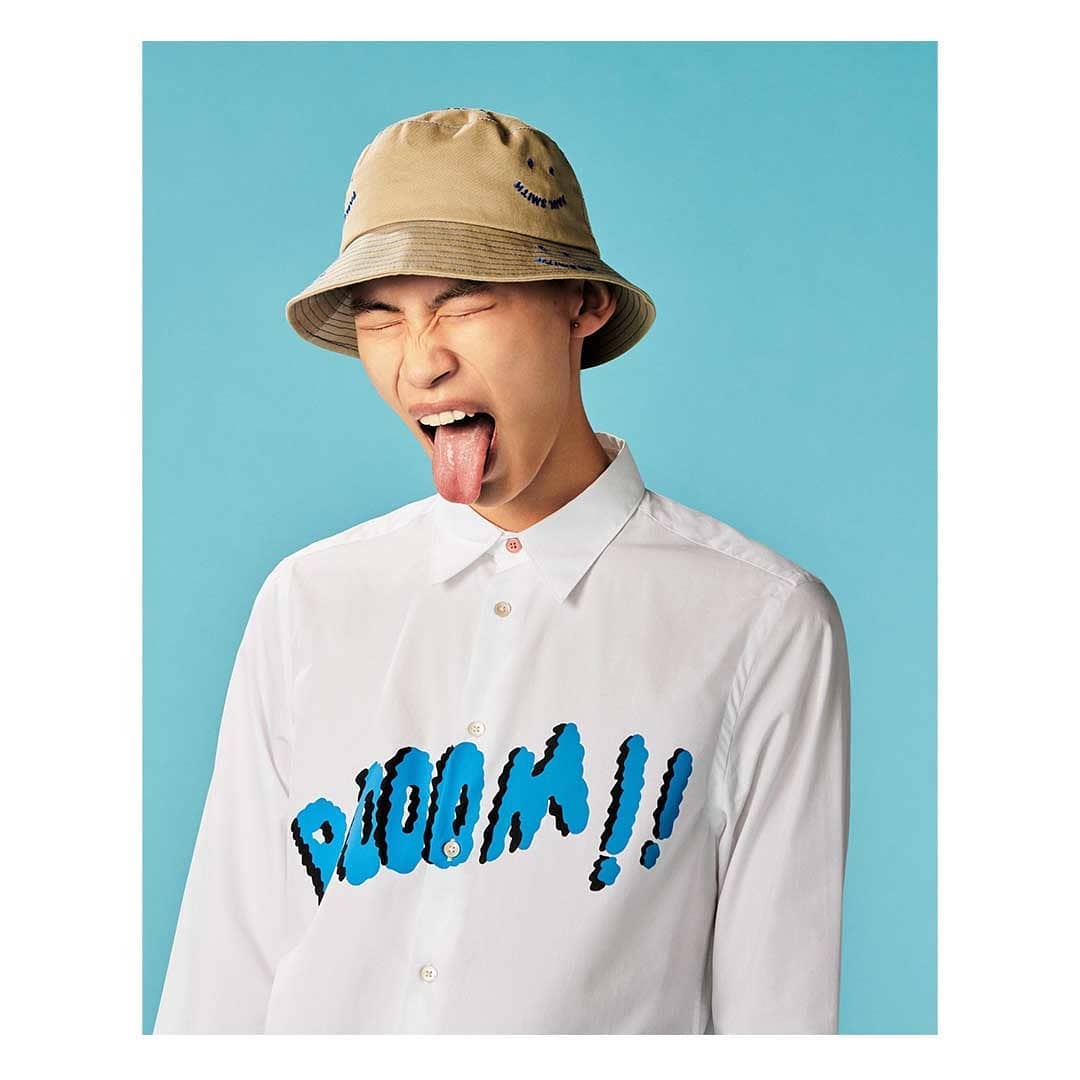 Asian man with tongue out wearing a tan bucket hat and white shirt with 'BOOOM!!' text against a blue background. Fashion editorial.