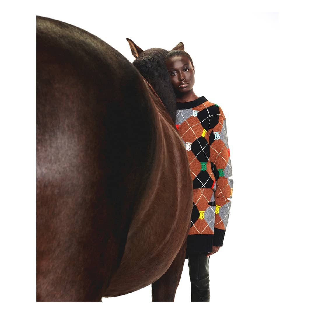 Fashion shot with a dark-skinned model wearing a sweater with an argyle pattern posing behind a dark brown horse in a bright studio.