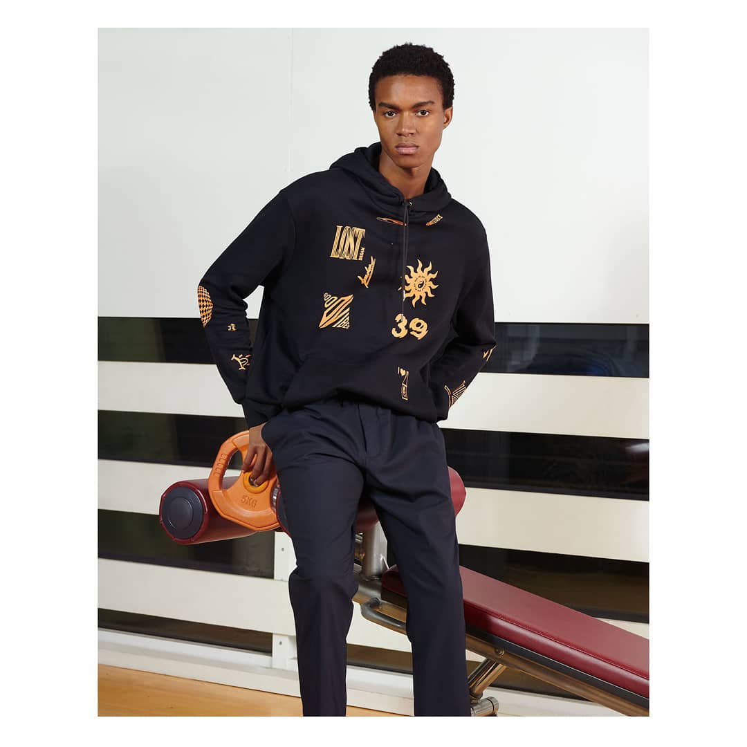 Adult male in black hoodie with graphics, navy pants, leaning on gym equipment in indoor setting. Fashion editorial photograph.