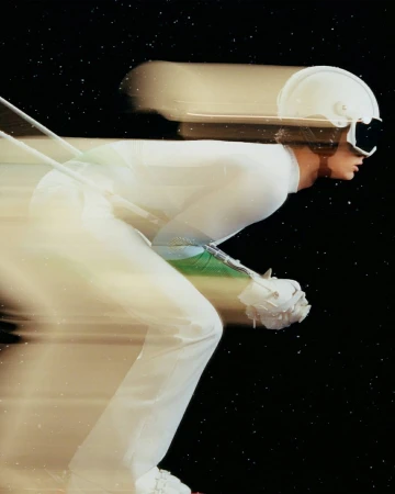 Motion blur photo of a person wearing a white ski suit, helmet, and goggles in a bent-over position, set against a dark background.