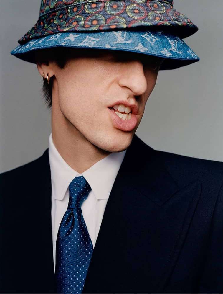 Close-up shot of a person wearing a colorful bucket hat and a navy suit with a blue patterned tie, set against a neutral background.