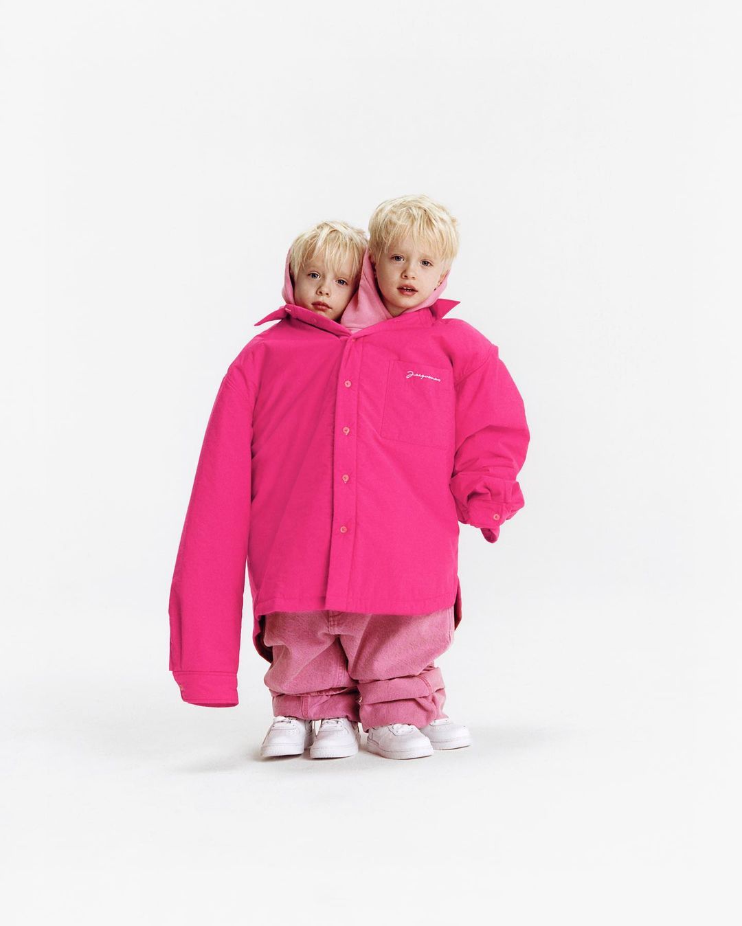 Two young children wearing oversized bright pink button-down shirt and matching pants and sneakers, posing against a white backdrop.