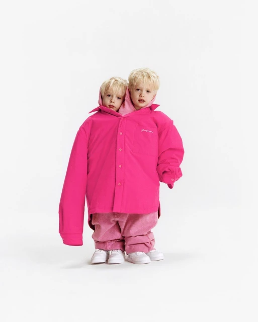 Two young children wearing oversized bright pink button-down shirt and matching pants and sneakers, posing against a white backdrop.