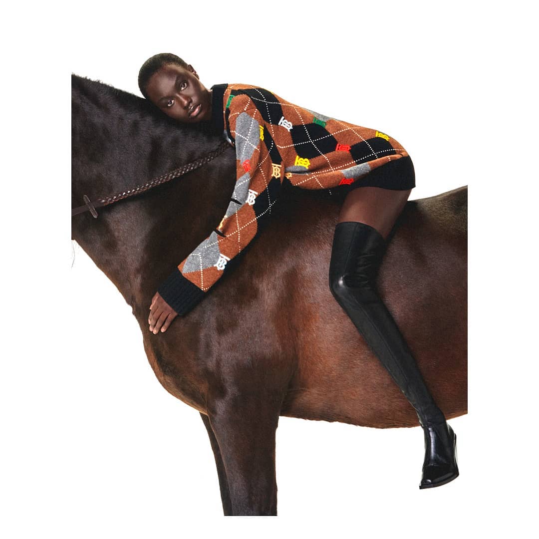 Adult woman in an argyle sweater and black thigh-high boots reclines on a brown horse in a fashion-style editorial shot.