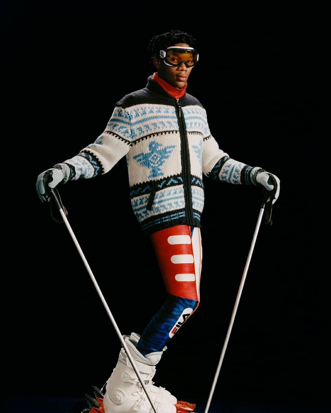 Fashion shot of African American male model in ski goggles, wearing an patterned sweater and ski pants while holding ski poles.