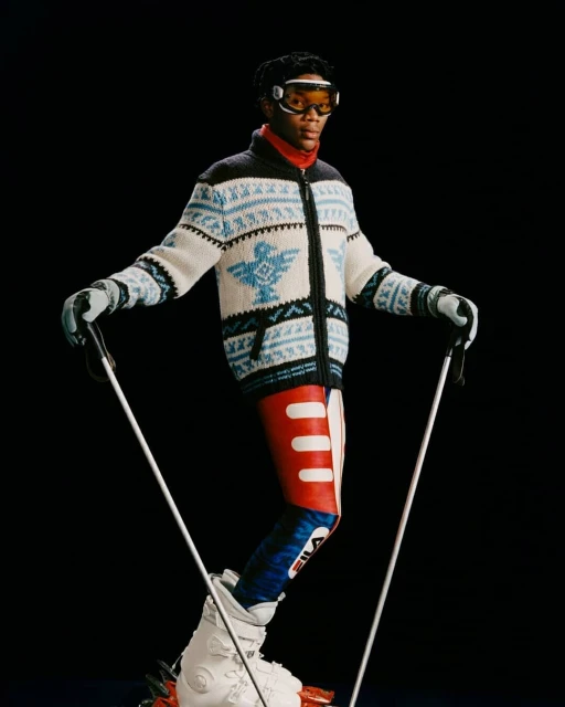 Fashion shot of African American male model in ski goggles, wearing an patterned sweater and ski pants while holding ski poles.