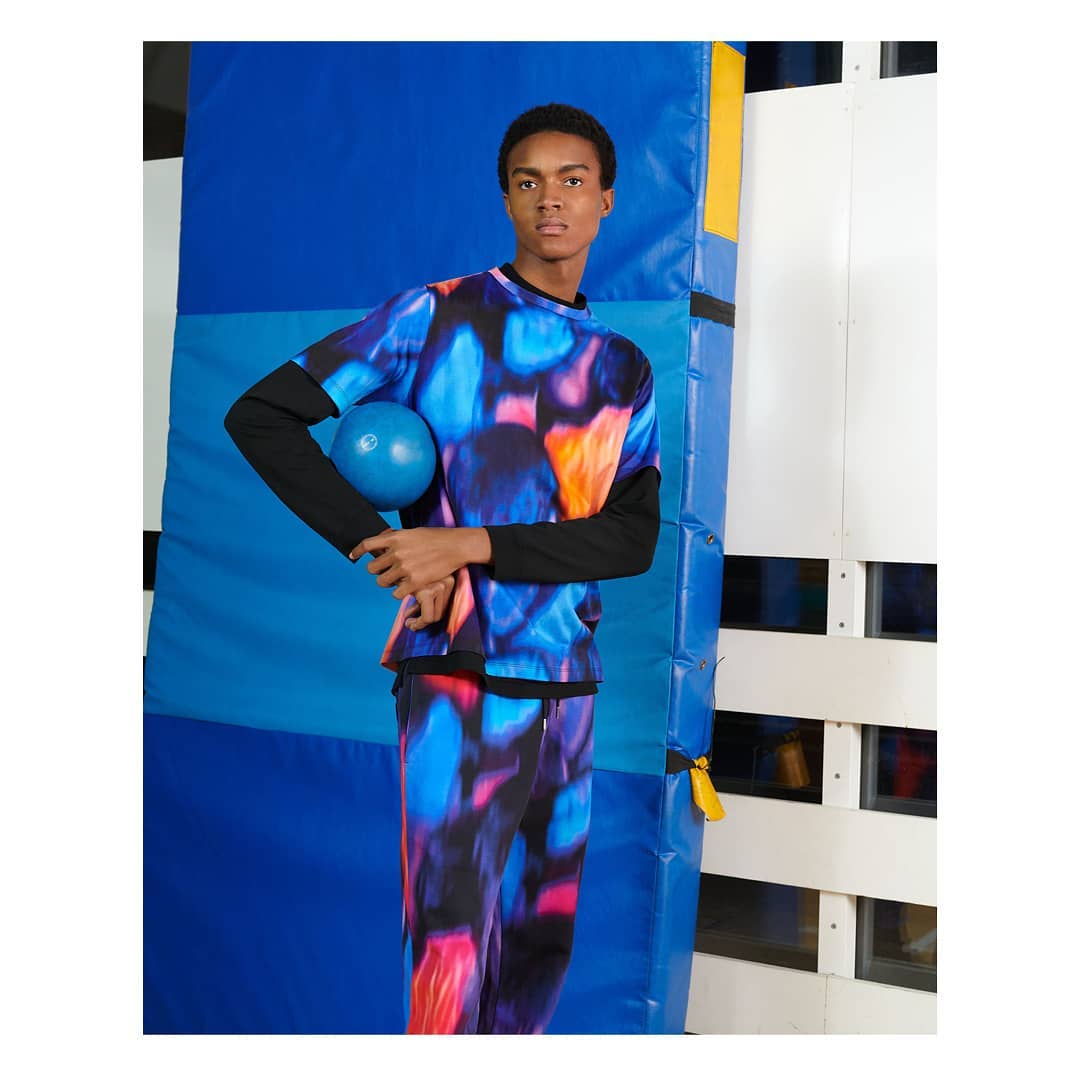 A dark-skinned male model in a colorful patterned top and pants holds a blue ball in a studio with blue and white wall accents.