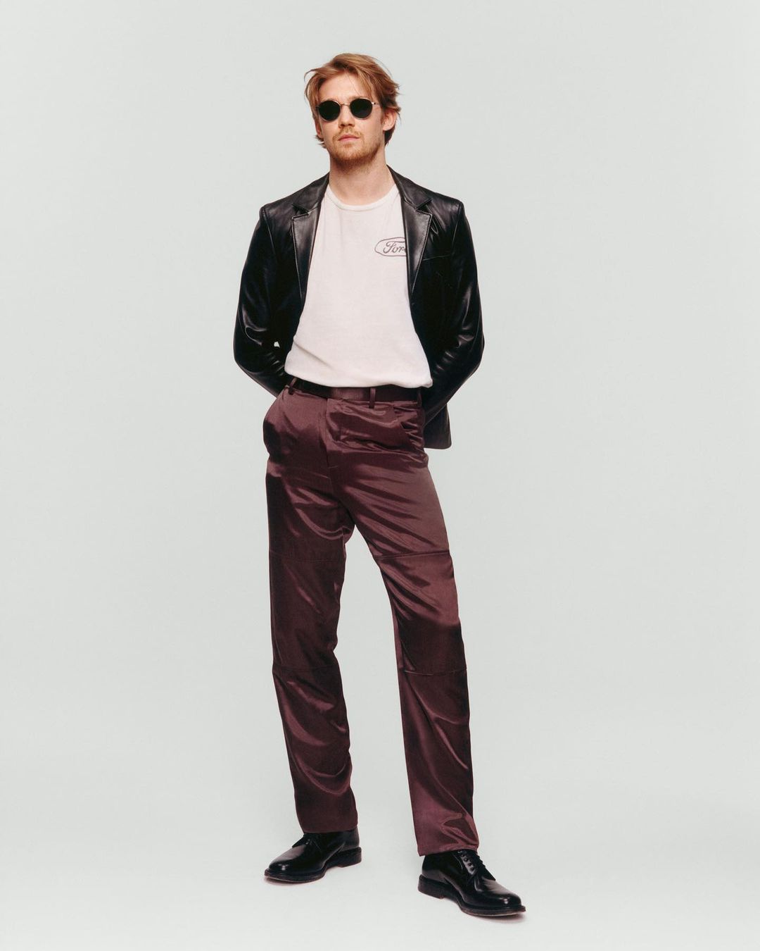 Man in sunglasses, leather jacket, t-shirt, and plum dress pants stands in a photo studio with neutral lighting and a solid background.