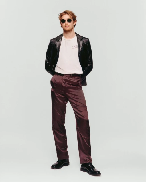 Man in sunglasses, leather jacket, t-shirt, and plum dress pants stands in a photo studio with neutral lighting and a solid background.