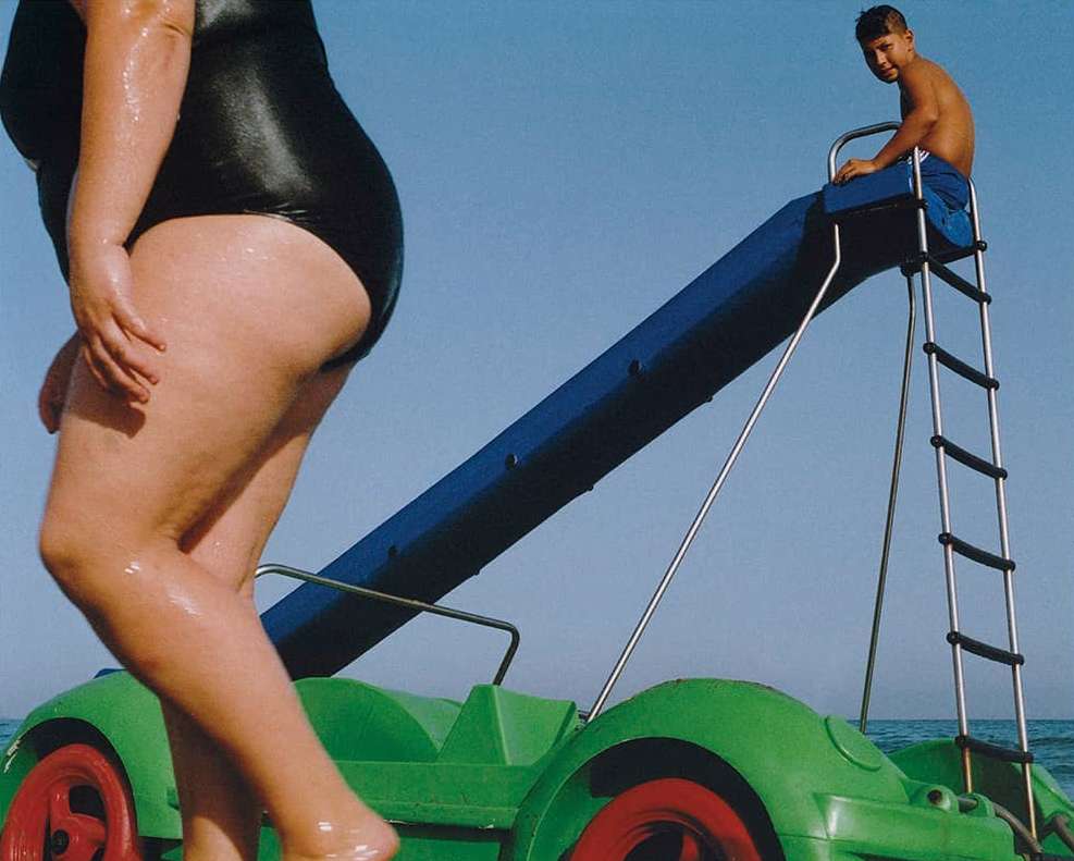 A woman stands near a blue water slide leading into the ocean while a boy is sitting at the top, posing for the photograph.