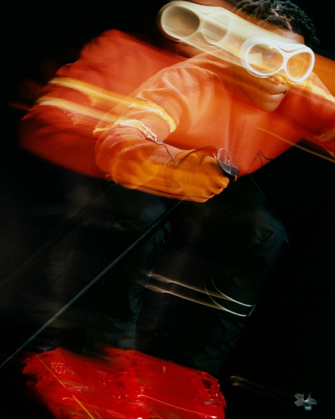 Blurred action shot of a person wearing glasses and an orange jacket, holding an object and moving against a dark background.