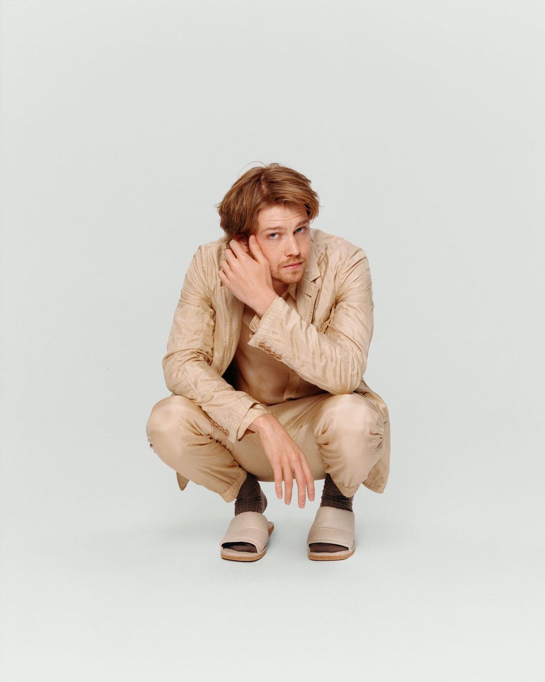 Adult male crouches in a photo studio wearing a tan suit and slides while touching his hair and glancing at the camera.