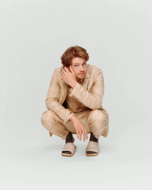 Adult male crouches in a photo studio wearing a tan suit and slides while touching his hair and glancing at the camera.