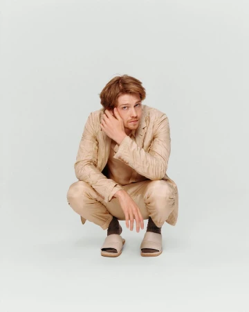 Adult male crouches in a photo studio wearing a tan suit and slides while touching his hair and glancing at the camera.