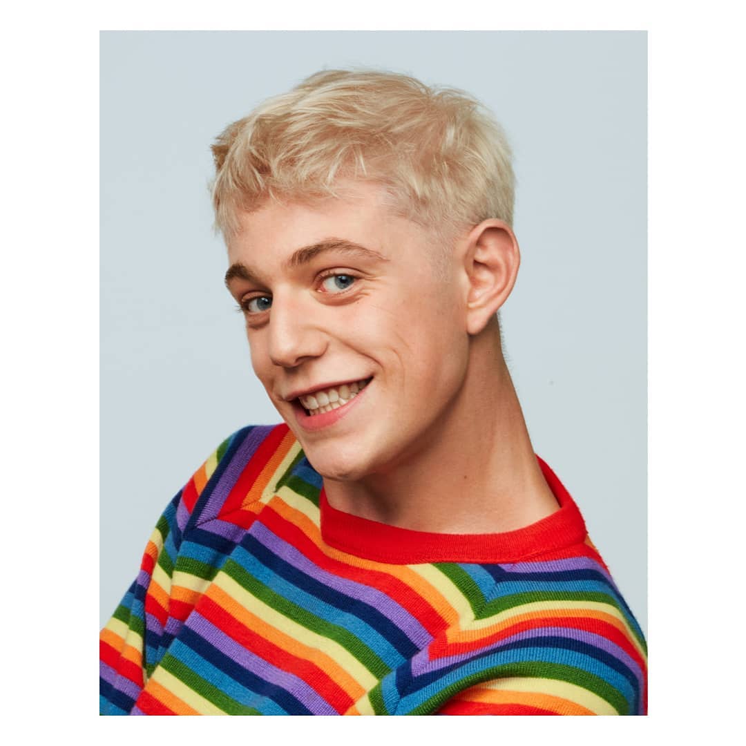 Portrait of a young adult with blonde hair smiling and wearing a colorful striped sweater against a solid light blue background.