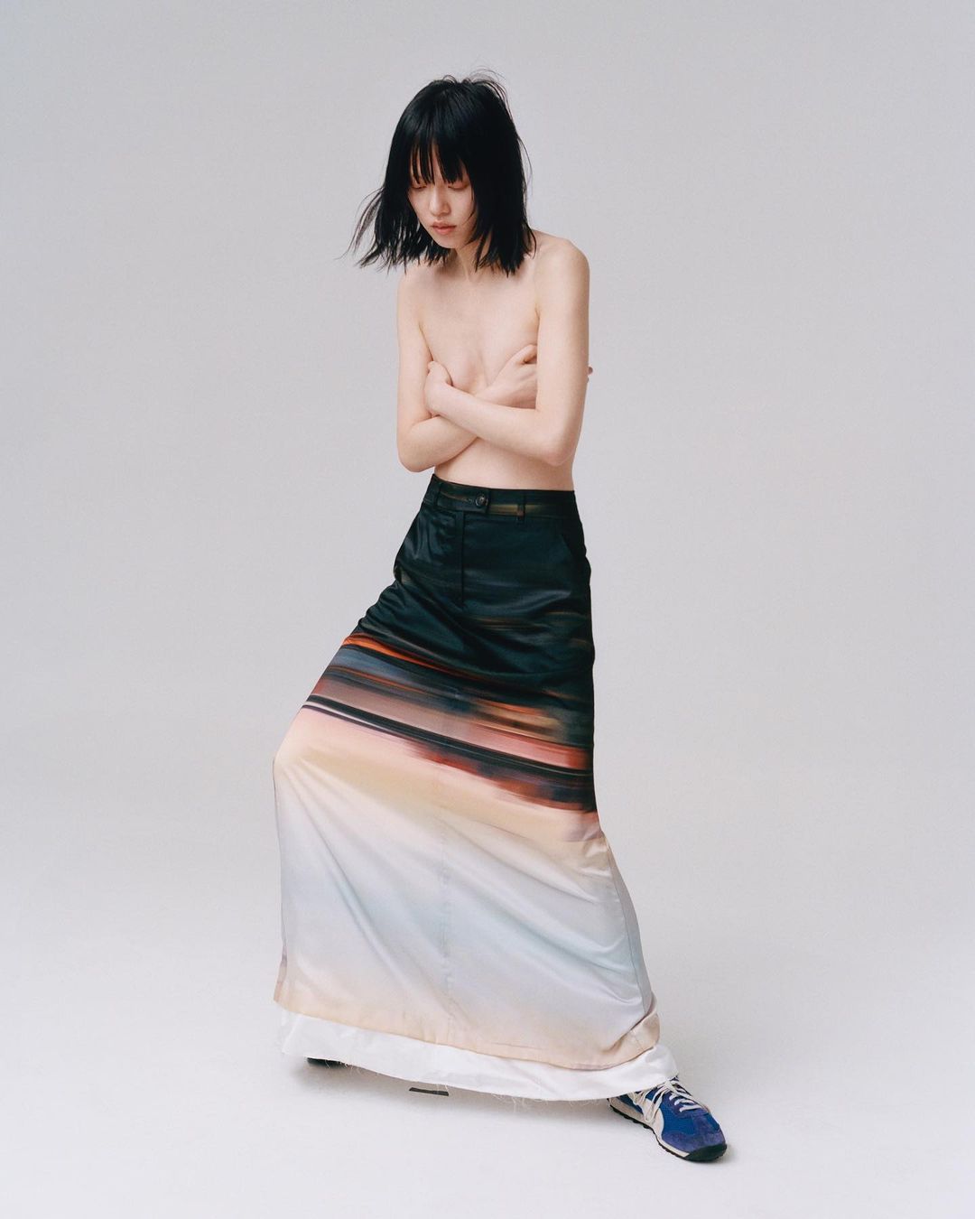 Bare-chested young woman with black hair, wearing a long skirt with a landscape print, poses in a bright studio.
