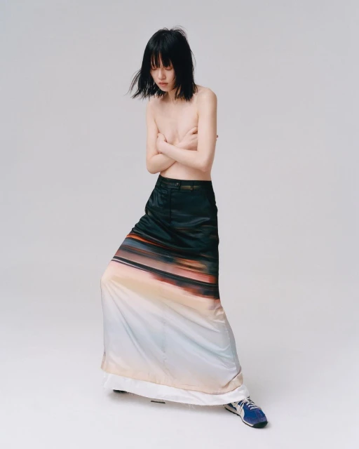 Bare-chested young woman with black hair, wearing a long skirt with a landscape print, poses in a bright studio.