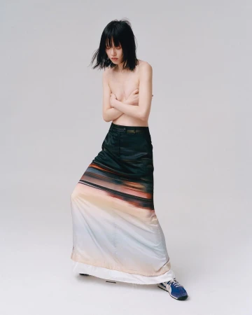 Bare-chested young woman with black hair, wearing a long skirt with a landscape print, poses in a bright studio.