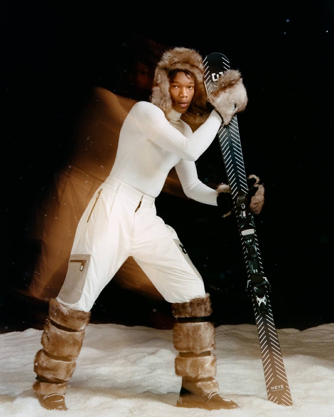 A person dressed in a white ski suit, wearing a fur hat, and holding skis in a snowy environment, against a dark background.