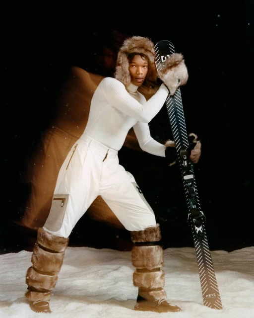 A person dressed in a white ski suit, wearing a fur hat, and holding skis in a snowy environment, against a dark background.