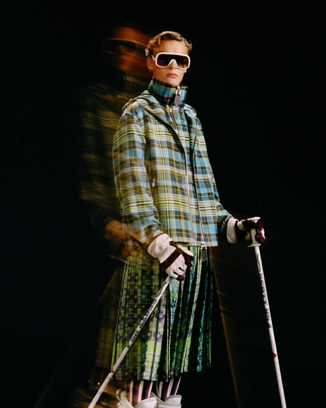 Person wearing a plaid jacket, pleated skirt, and ski goggles holds ski poles against a dark background in a studio fashion shot.