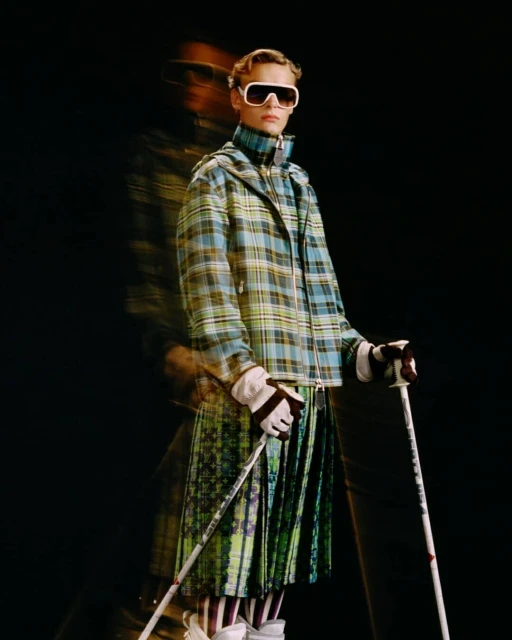 Person wearing a plaid jacket, pleated skirt, and ski goggles holds ski poles against a dark background in a studio fashion shot.