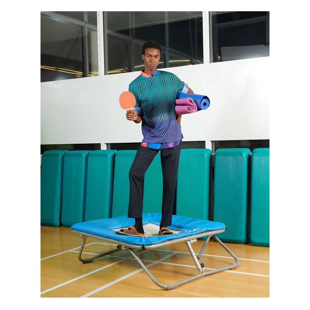 Athletic man in patterned shirt and dark pants holding ping pong paddle and yoga mats stands on small trampoline indoors.