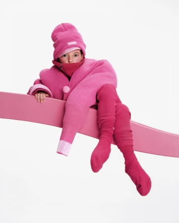 Young child wearing a pink hat, sweater, scarf, leggings, and socks, sitting on a pink bar in a brightly lit, white studio.