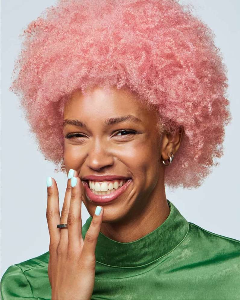 Close-up of a smiling Black woman with a large, pink afro, wearing a green high-necked satin top. She is holding up her hand near her face.