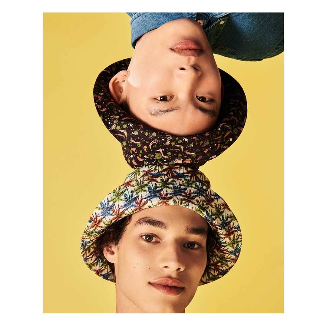 Two young adult men pose against a vibrant yellow background, each wearing a distinct bucket hat in a studio portrait.