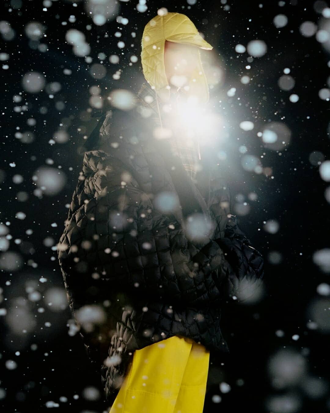 A person in a black quilted jacket and yellow pants stands in front of a black background with bokeh effect, creating a dreamy, wintery vibe.