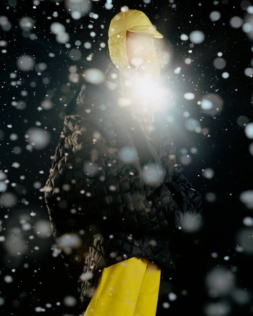 A person in a black quilted jacket and yellow pants stands in front of a black background with bokeh effect, creating a dreamy, wintery vibe.