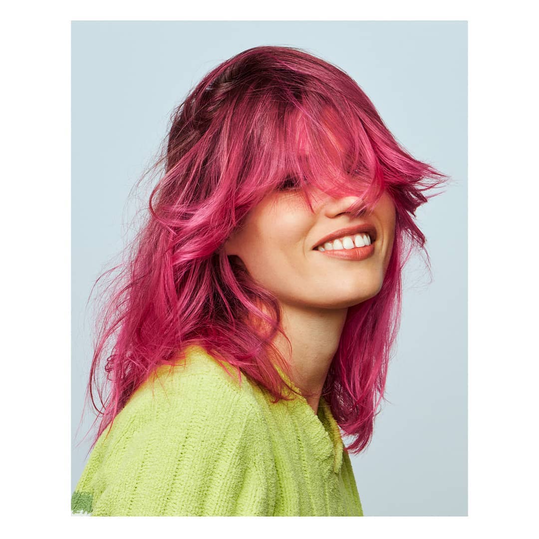 Smiling woman with dyed pink hair and light green knit sweater against a light blue background in a medium studio shot.