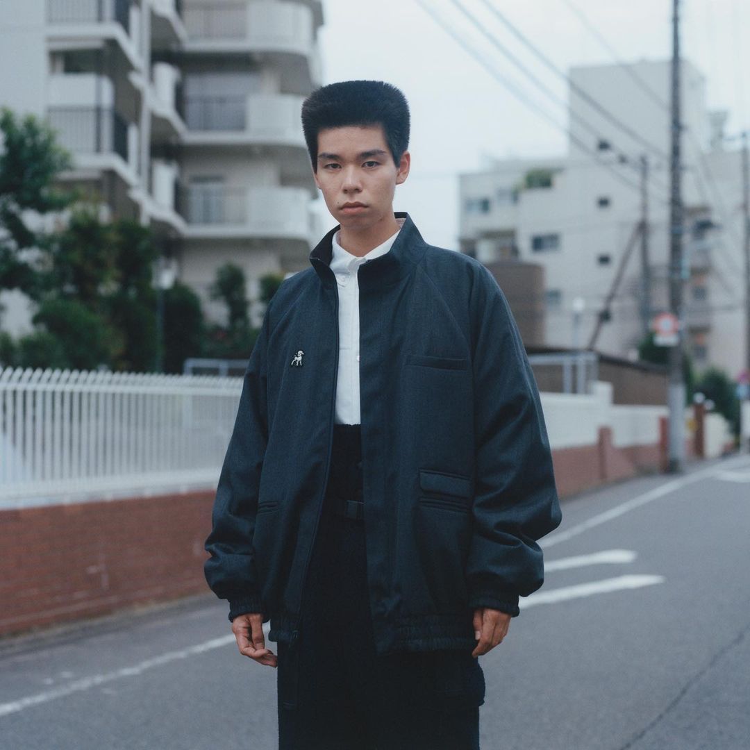 Portrait of a young adult in a dark jacket and black pants stands on an asphalt street, with apartment buildings visible in the background.