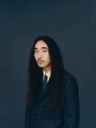 Portrait of a man with long dark hair and mustache wearing a dark pinstripe suit jacket photographed by Fumi Homma