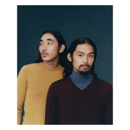 Two men with long dark hair and facial hair posing against a dark studio background in London photographed by Fumi Homma