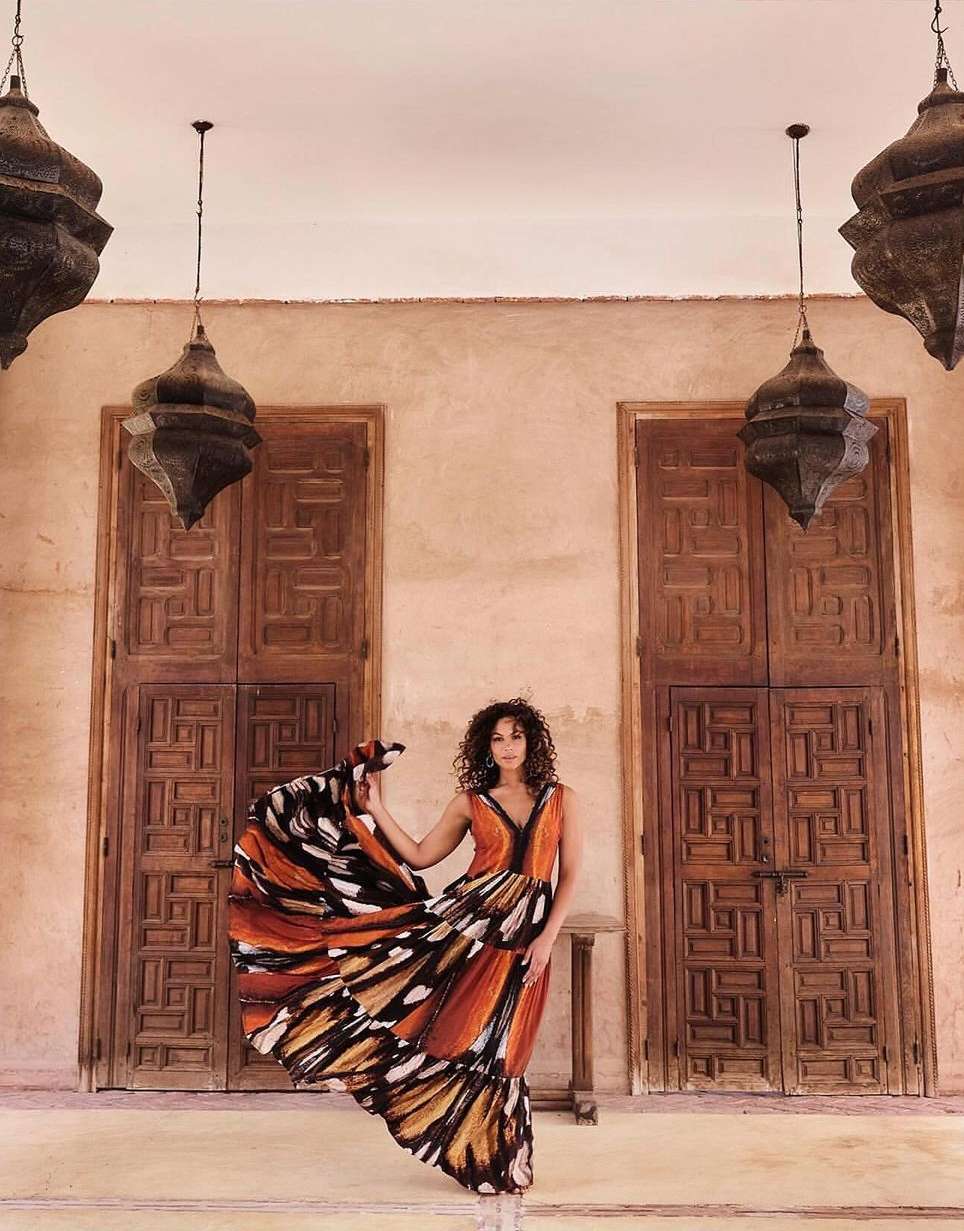 Woman in long butterfly-print dress poses in Moroccan building with decorative lamps, wooden doors, and neutral-toned walls and floor.