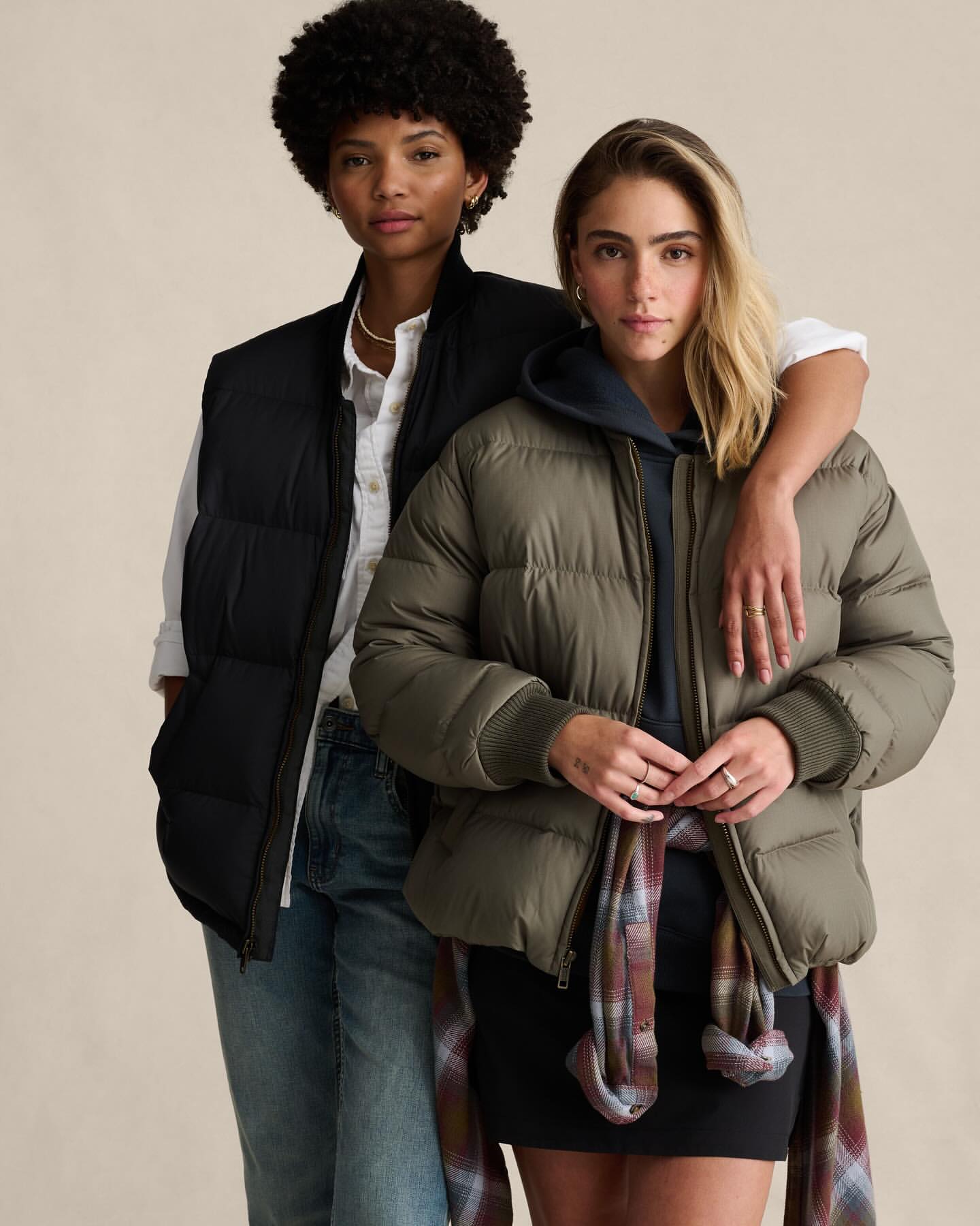 Two women pose indoors in winter clothing. One wears a black puffer vest and jeans; the other a khaki jacket, short skirt, and plaid scarf.