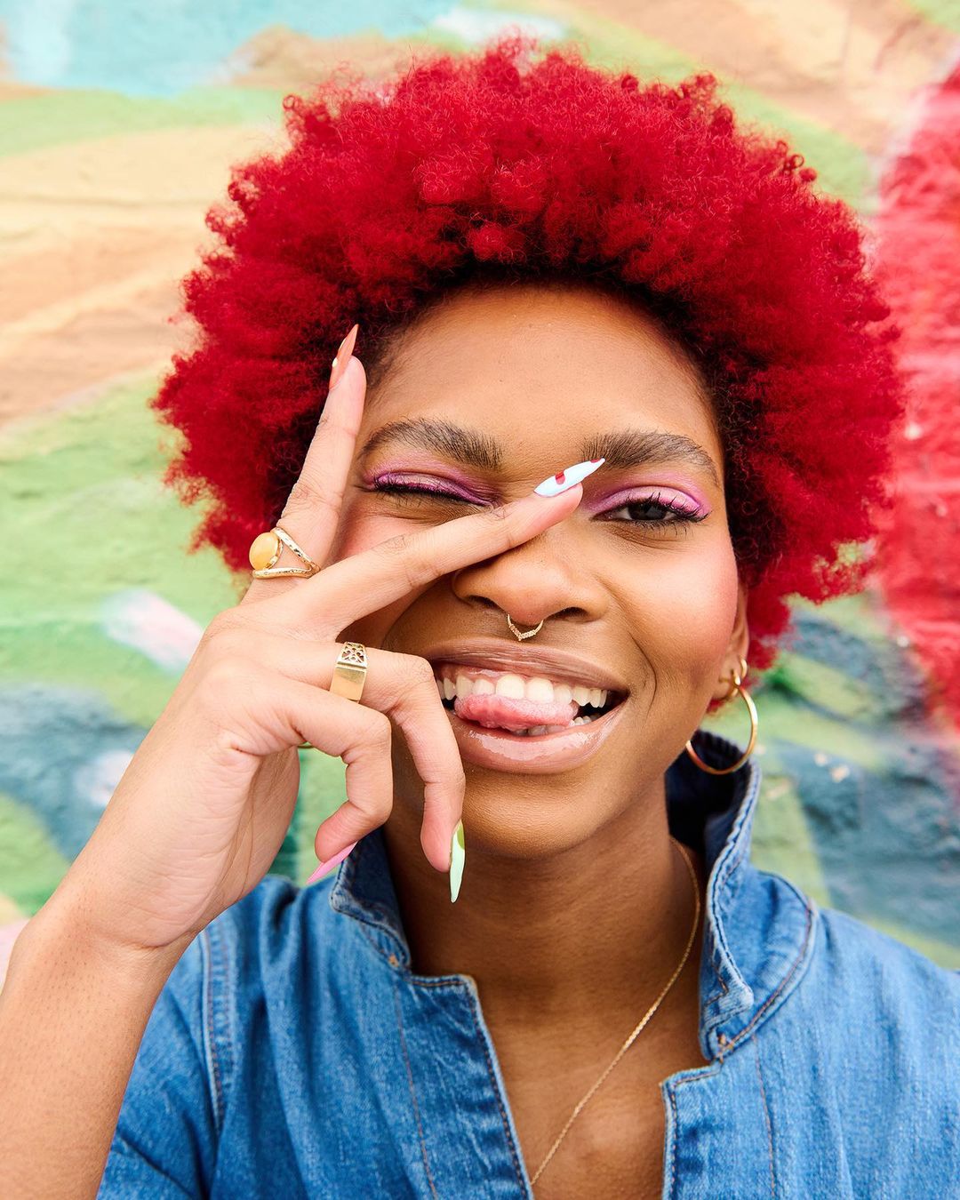 Portrait of an adult woman with red curly hair winking, wearing a denim jacket and gold jewelry, with a colorful, blurry background.