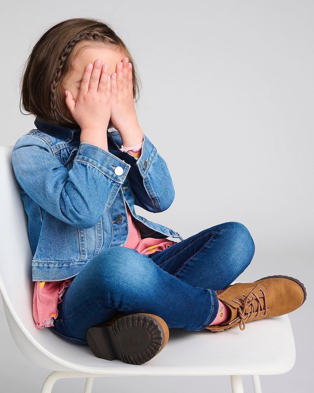 Young child covers her face while seated on a white chair indoors. She wears a denim jacket, jeans, and brown boots in a studio setting.